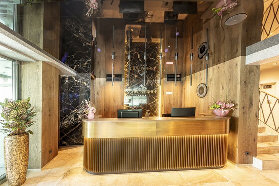 ©MartinRaffeiner_07_2024_RF_5349 Ein moderner Empfangsbereich mit einem eleganten, goldenen Empfangstresen. Die Wände sind aus Holz und Stein, dekoriert mit Pflanzen und stilvollen Beleuchtungselementen.