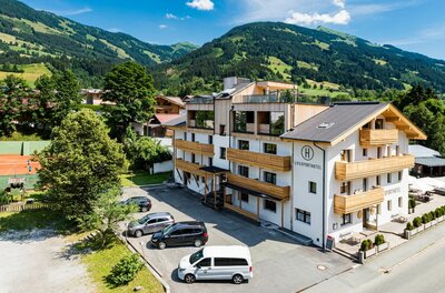 Mehrstöckiges Hotelgebäude mit Holzbalkonen vor bewaldeten Bergen und einem Parkplatz im Vordergrund.