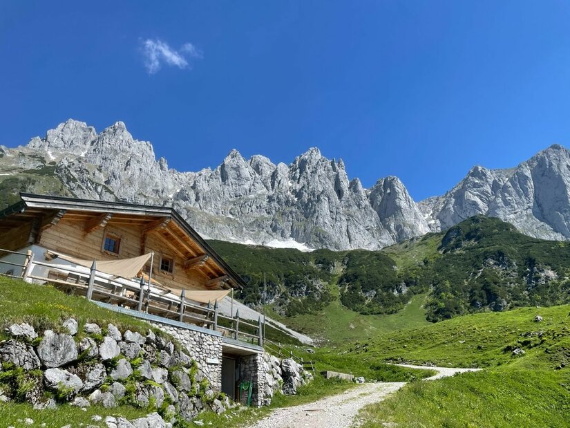 Obere Regalm vor Bergmassiv Holzhütte auf einem Grashang vor einem schroffen Felsmassiv unter blauem Himmel.