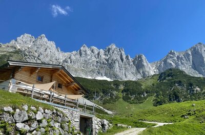 Holzhütte auf einem Grashang vor einem schroffen Felsmassiv unter blauem Himmel.
