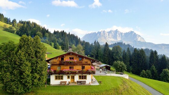 Traditionelles Holzhaus mit Blumenschmuck vor bewaldeten Hügeln und dem Wilden Kaiser Gebirge.
