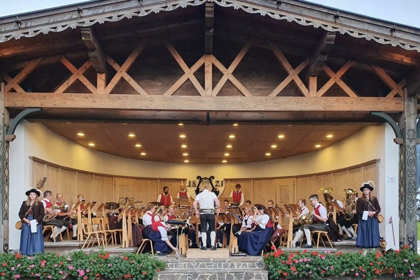 Eine festliche Veranstaltung mit einer Musikgruppe auf einer Bühne. Die Musiker tragen traditionelle Kleidung und umgeben sich mit Stühlen und Blumen.