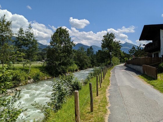 Weg am Fluss in Malerndörfl Asphaltierter Weg neben einem fließenden Fluss mit Bergen im Hintergrund.