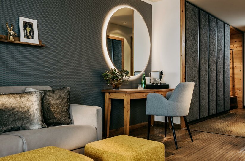 Zimmer im Grand Tirolia Modernes Hotelzimmer mit Sofa, Holztisch, beleuchtetem Rundspiegel und grauem Sessel.
