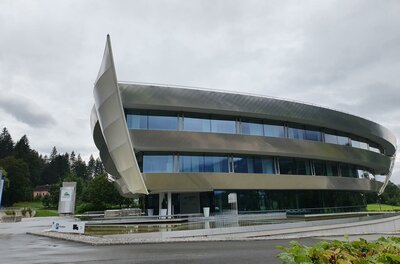 Ein modernes, futuristisch gestaltetes Gebäude mit großen Fenstern und einer geschwungenen Fassadenstruktur. Es liegt in einer grünen Umgebung unter einem bewölkten Himmel.