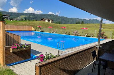 Schwimmbad Itter (c)Clarissa Günnewig Ein schwimmendes, klares blaues Wasserbecken mit Blumen und Liegestühlen. Im Hintergrund sind grüne Wiesen und Berge sichtbar.