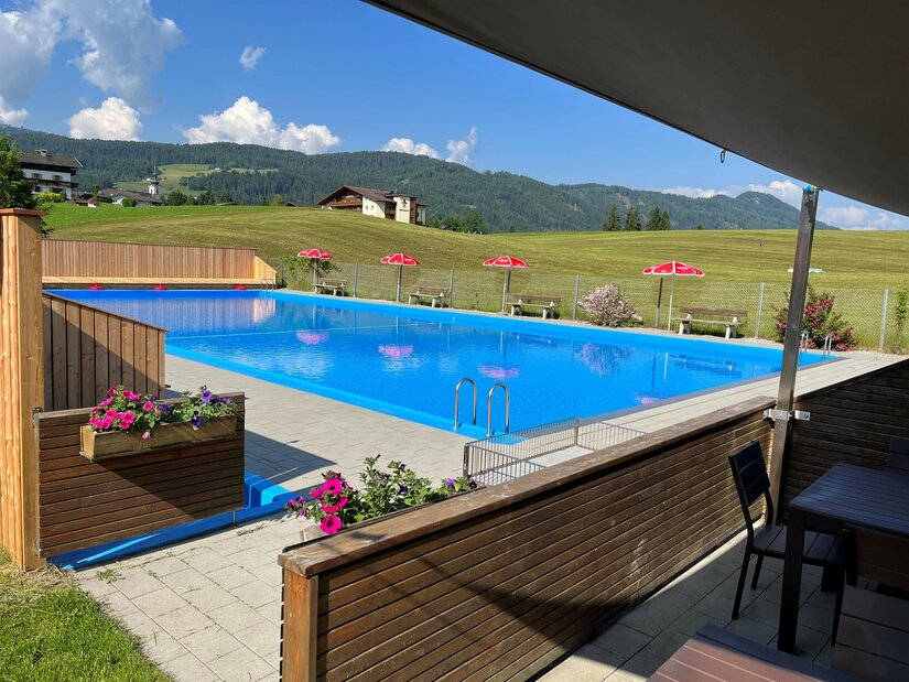 Schwimmbad Itter (c)Clarissa Günnewig Ein schwimmendes, klares blaues Wasserbecken mit Blumen und Liegestühlen. Im Hintergrund sind grüne Wiesen und Berge sichtbar.