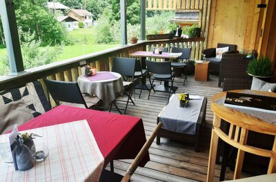 Eine gemütliche Terrasse mit Tischen und Stühlen. Im Hintergrund sieht man eine grüne Landschaft und einige Gebäude.