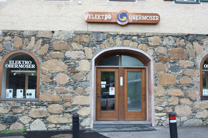 Ein Gebäudeeingang mit zwei Holz-Türen und einem Schild mit der Aufschrift "Elektro Obermoser". Die Fassade ist aus Steinen und weist zwei Fenster auf.