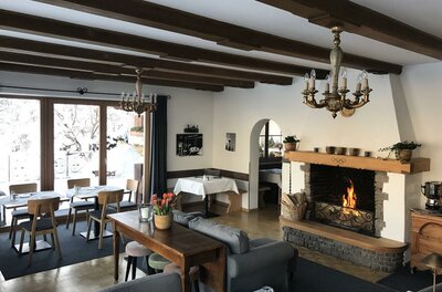 Kaminzimmer der Pension Hinterseer Innenansicht eines Gastraums mit brennendem Kamin, Holzbalkendecke und eingedeckten Tischen.