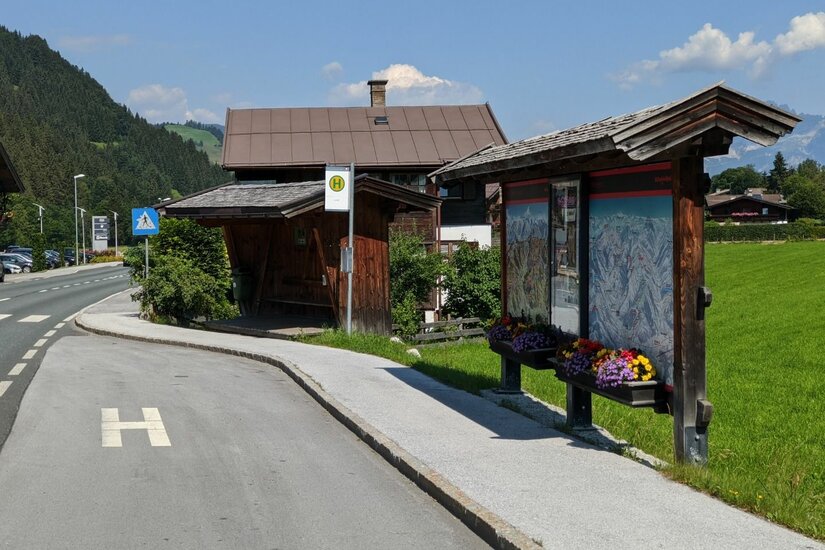 Bushaltestelle mit Holzunterstand und Informationstafel an einer Straße vor Bergkulisse.