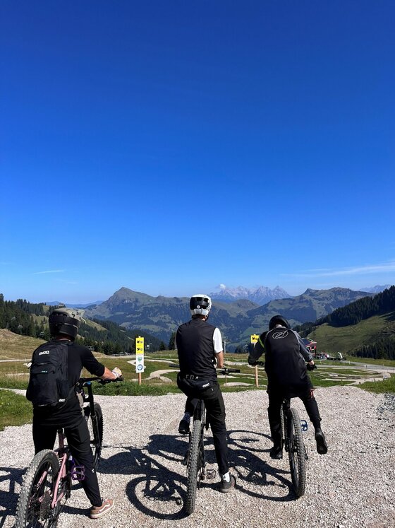 Mountainbiker an der Family Area Drei Mountainbiker stehen mit ihren Rädern auf einem Schotterweg vor einem Bergpanorama unter blauem Himmel.