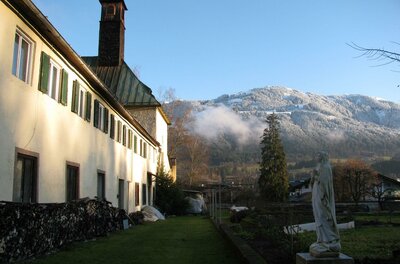 Kloster Ein charmantes Gebäude mit grünen Fensterläden und einem blauen Himmel im Hintergrund. Im Vordergrund steht eine Statue und die Umgebung ist von schneebedeckten Bergen umgeben.