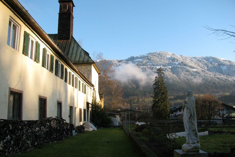 Ein charmantes Gebäude mit grünen Fensterläden und einem blauen Himmel im Hintergrund. Im Vordergrund steht eine Statue und die Umgebung ist von schneebedeckten Bergen umgeben.