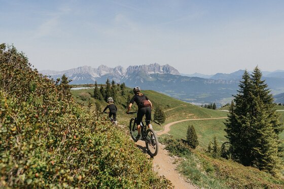Mountainbiken am Hahnenkamm Trail Zwei Mountainbiker fahren auf einem schmalen Pfad bergab vor einem Gebirgspanorama mit schroffen Felsgipfeln.