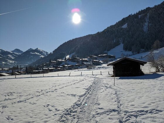 Verschneite Wiese mit Spuren im Schnee, einer Holzhütte und einem Dorf vor bewaldeten Bergen unter blauem Himmel.