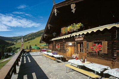 Sonnenterrasse einer hölzernen Almhütte mit Sitzbänken und Blick auf grüne Berghänge.