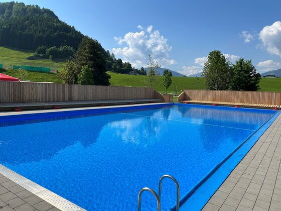 Ein großer, blauer Swimmingpool umgeben von einer Holzumrandung und grünen Wiesen. Im Hintergrund sind sanfte Hügel und ein klarer Himmel zu sehen.