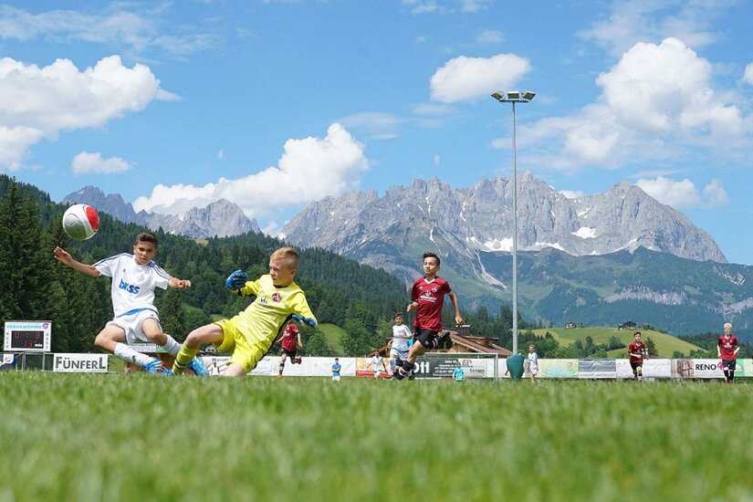 Jugendliche beim Fußballspiel auf einem Rasenplatz vor einer alpinen Bergkulisse mit Felsmassiv.