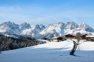 Verschneite Almhütten vor einem massiven, schneebedeckten Gebirgszug unter blauem Himmel.