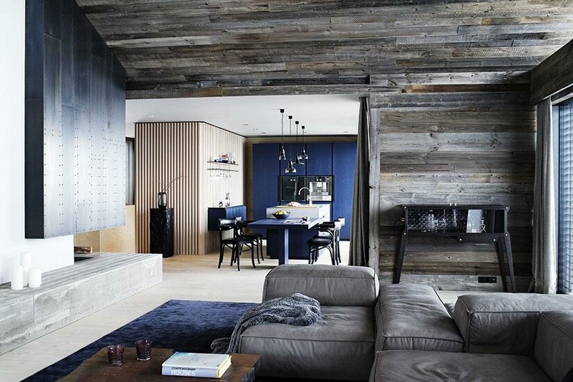 Moderner alpiner Wohnbereich Moderner Wohnraum mit Altholzwänden, grauem Sofa und blauer Küche im Hintergrund.