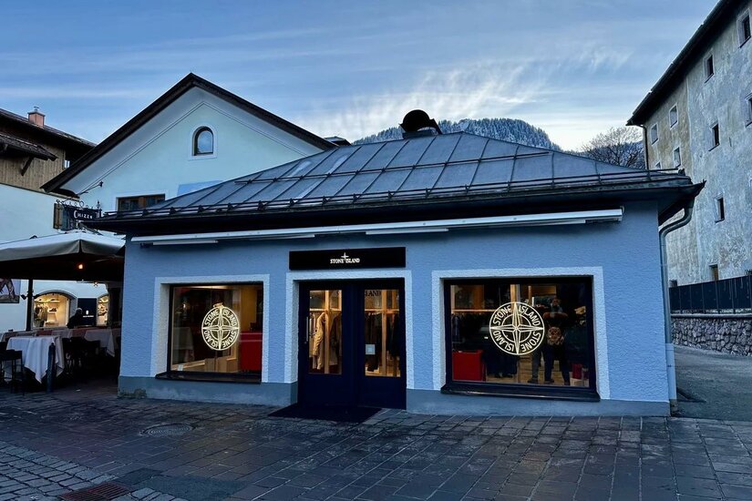Stone Island Store Außenansicht eines Stone Island Ladengeschäfts in einem hellblauen Gebäude mit Bergpanorama im Hintergrund.