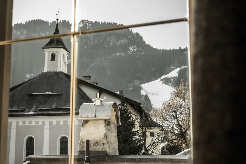 Ein Blick aus einem Fenster auf ein malerisches Dorf mit einer Kirche. Im Hintergrund sind schneebedeckte Berge zu sehen.