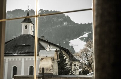 Apart_Mantinger_Kitz_Klammer-07736 Ein Blick aus einem Fenster auf ein malerisches Dorf mit einer Kirche. Im Hintergrund sind schneebedeckte Berge zu sehen.