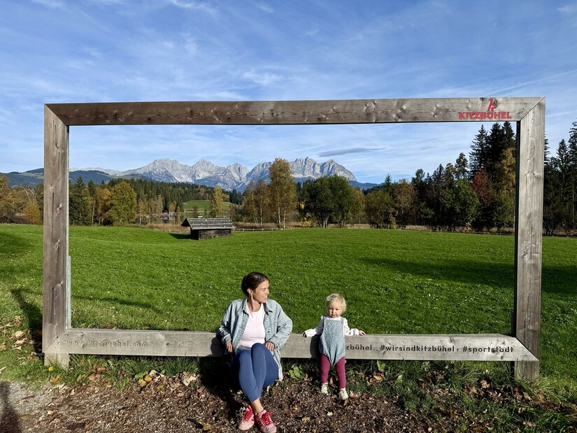 Frau und Kleinkind sitzen in einem großen Holzrahmen vor einer Bergkulisse und grünen Wiesen.