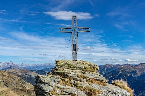 Gipfelkreuz am Tristkogel Ein metallisches Gipfelkreuz auf einem felsigen Berggipfel vor blauem Himmel und Gebirgspanorama.
