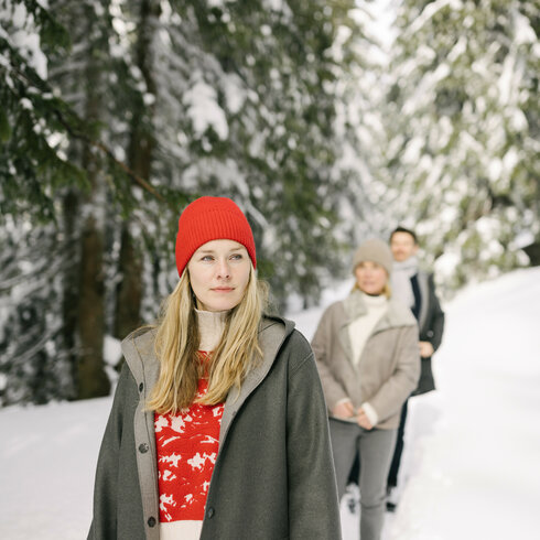 Hinter einer Frau mit rotem Hut und langen Haaren sind drei Personen zu sehen, die in einem verschneiten Wald spazieren gehen.