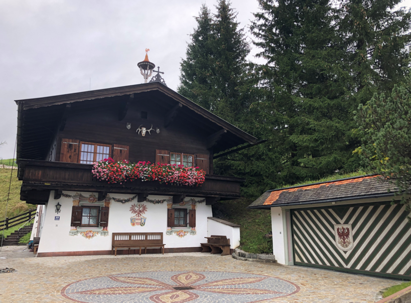 Chalet Marie-Luise Traditionelles Alpenhaus mit dunkler Holzfassade, Blumenbalkon und Wandmalereien vor Nadelbäumen.