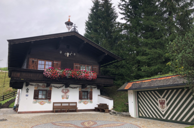 Traditionelles Alpenhaus mit dunkler Holzfassade, Blumenbalkon und Wandmalereien vor Nadelbäumen.
