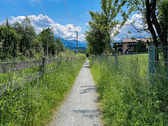 Stadtrunde Schotterweg Schmaler Schotterweg zwischen Holzzaun und Metallzaun mit Blick auf Berge.