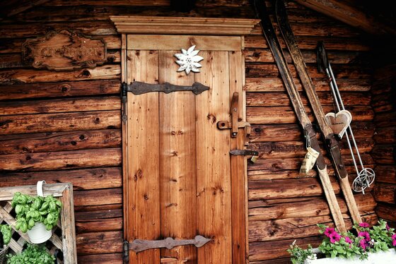 Eingangsbereich Astlingerhof Holztür einer Almhütte mit Edelweiß-Verzierung, daneben lehnen alte Holzski an der Blockwand.