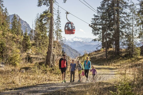 Wandern auf der Steinplatte Eine Familie wandert auf einem Weg in den Bergen. Im Hintergrund ist eine Seilbahn und schneebedeckte Gipfel zu sehen.