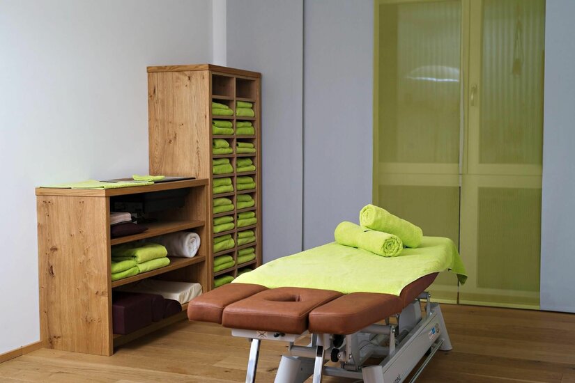 Behandlungsraum Praxis Wilder Kaiser Eine braune Massageliege mit grünen Handtüchern vor einem Holzregal in einem hellen Behandlungsraum.
