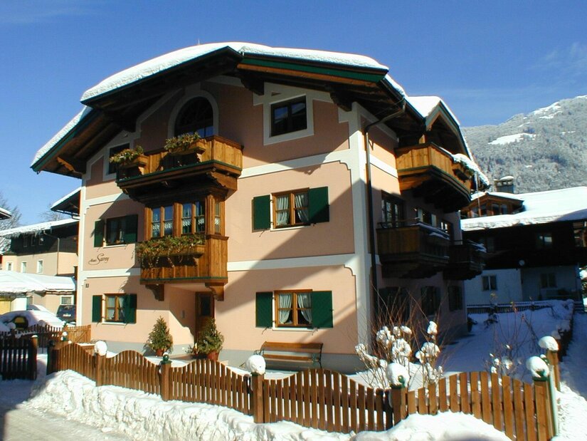 Mehrstöckiges alpines Wohnhaus mit Holzfassadenelementen und schneebedecktem Dach unter blauem Himmel.