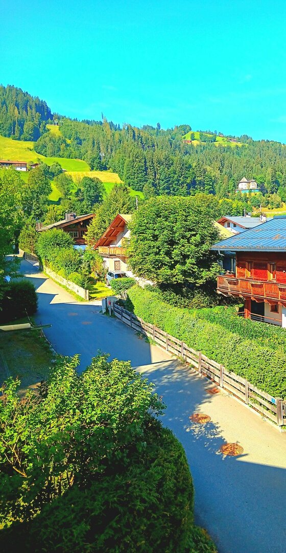 Blick zum Hahnenkamm Blick von oben auf eine asphaltierte Straße, gesäumt von Holzhäusern und grünen Hecken vor bewaldeten Bergen.