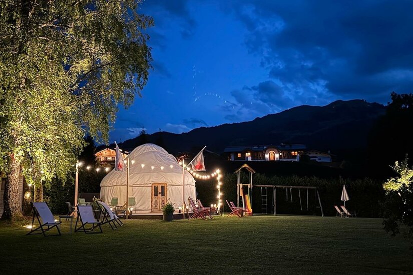 Beleuchtete weiße Jurte auf einer Wiese bei Nacht mit Liegestühlen und Bergen im Hintergrund.