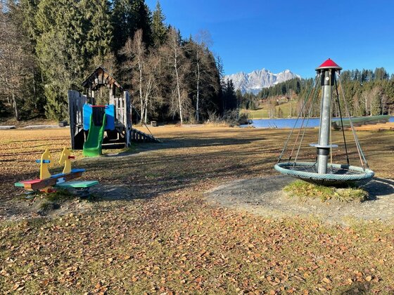 Kinderspielplatz auf einer Wiese mit Rutsche, Wippe und Karussell vor einem See und Bergen.