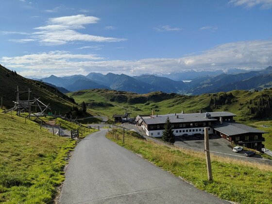 Berggasthof am KAT Walk Asphaltierter Wanderweg führt bergab zu einem großen Berggasthof in einer alpinen Wiesenlandschaft mit Bergpanorama.