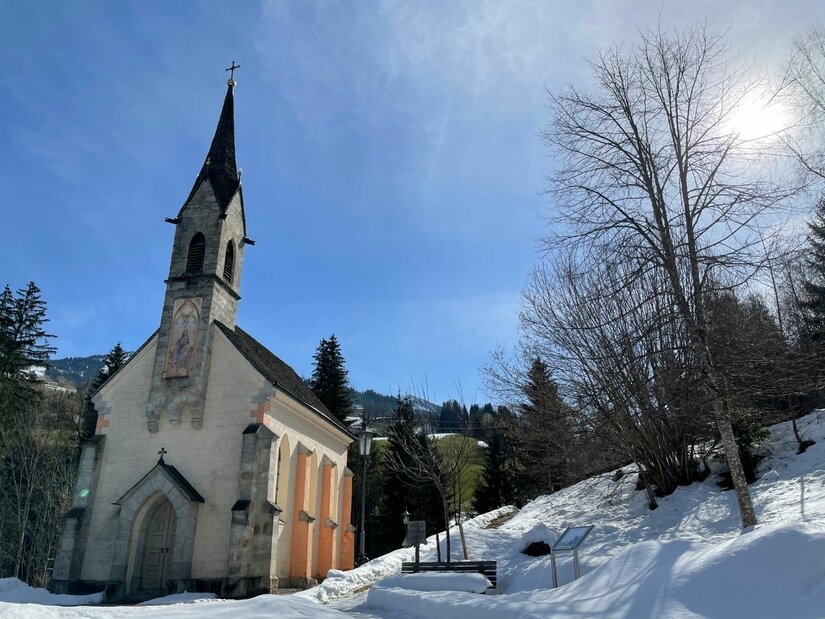 Kleine Kapelle mit spitzem Turm in einer verschneiten Winterlandschaft unter blauem Himmel.