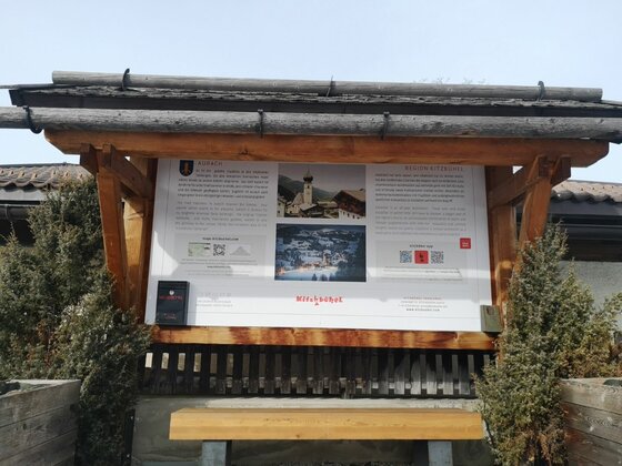 Infotafel am Achenweg Aurach Informationstafel aus Holz mit Dach und Sitzbank vor Nadelgehölzen.