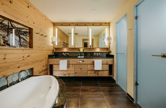 Badezimmer im Grand Tirolia Modernes Badezimmer mit Holzverkleidung, Doppelwaschbecken und einer weißen Badewanne im Vordergrund.