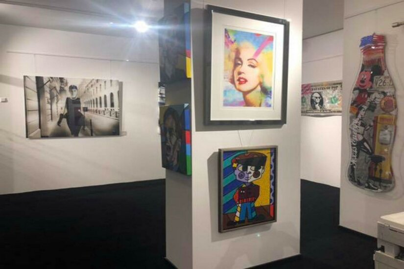 Walentowski Gallerie Jochberg Eine Kunstgalerie mit mehreren bunten Gemälden und Fotografien an den Wänden. Der Boden ist schwarz und die Beleuchtung ist hell und freundlich.