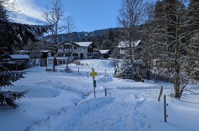 Winterwanderweg in Hörpfing Verschneiter Wanderweg führt zu einem gelben Wegweiser vor alpinen Häusern und Nadelbäumen.
