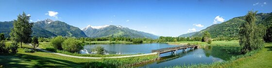 Panorama eines Sees mit Holzbrücke, umgeben von Wiesen und Bergen unter blauem Himmel.