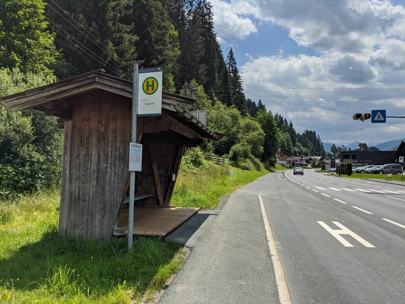 Hölzernes Wartehäuschen einer Bushaltestelle an einer Landstraße neben einem Wald.