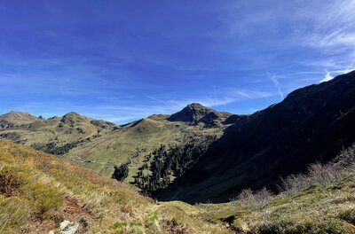 Panorama am Mittagskogel Blick von einem grasbewachsenen Berghang auf gegenüberliegende Berggipfel unter blauem Himmel.